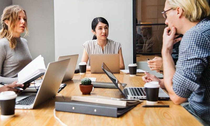 A group of professionals engaged in a meeting around a wooden table with laptops, notepads, and coffee cups.