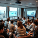 A group of professionals engages in a sales training in London, and discussing data on large screens with a cityscape view from a high-rise office.